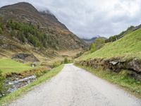 a gravel road running through a green valley with a stream in between two cliffs and grass on either side of the road