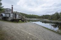 an old train carriage sitting in front of a pond near the road and trees on both sides and steps