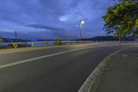 a road with a street light on the side near a lake and sky filled with clouds