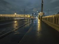 a wet sidewalk next to a road with buildings on it at night with train tracks in foreground