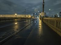 a wet sidewalk next to a road with buildings on it at night with train tracks in foreground