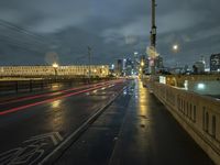 a wet sidewalk next to a road with buildings on it at night with train tracks in foreground