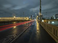 a wet sidewalk next to a road with buildings on it at night with train tracks in foreground