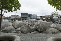 large rocks in the street near several buildings and trees are surrounded by green leaves on a cloudy day