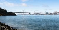 the suspension bridge over the water next to a rocky shore and a large city in the background