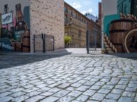 a gate is seen at the end of the street with graffiti on it, there is no one walking on the brick street or in front