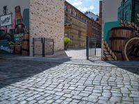 a gate is seen at the end of the street with graffiti on it, there is no one walking on the brick street or in front