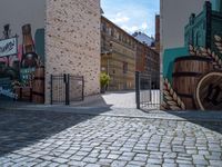 a gate is seen at the end of the street with graffiti on it, there is no one walking on the brick street or in front