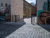 a gate is seen at the end of the street with graffiti on it, there is no one walking on the brick street or in front