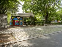 a mural on a stone wall at a bus stop with graffiti covering it in front of trees