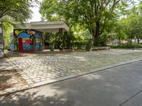 a mural on a stone wall at a bus stop with graffiti covering it in front of trees