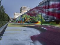 the long train has graffiti on the side of it on a street corner with tall buildings in the background