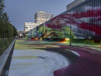 the long train has graffiti on the side of it on a street corner with tall buildings in the background