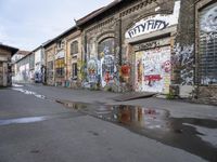 graffiti in a street with wet pavement and buildings with signs on them and a puddle