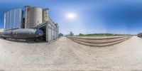 a fish eye view of a train at a factory with oil tanks behind it and the sun in the background