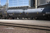 a black train parked next to a group of tanks next to a building and a lot of tracks