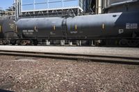 a black train parked next to a group of tanks next to a building and a lot of tracks