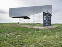 a glass structure with wind turbines behind it, on a grassy hill in front of gray building