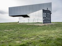 a glass structure with wind turbines behind it, on a grassy hill in front of gray building