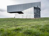 a glass structure with wind turbines behind it, on a grassy hill in front of gray building