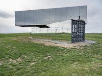 a glass structure with wind turbines behind it, on a grassy hill in front of gray building