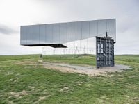 a glass structure with wind turbines behind it, on a grassy hill in front of gray building