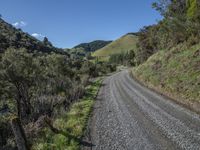 a gravel road winding down through grassy green mountains with blue skies in the distance at a glance