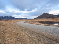 a gravel road with mountains in the background of the photograph, a lake and grass are below