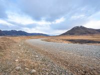 a gravel road with mountains in the background of the photograph, a lake and grass are below