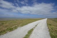 a gravel road leading to an ocean next to a dirt field on a cloudy day