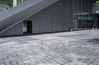 an image of a gray building in the city with a stairs outside the door and on a brick area with the buildings behind it