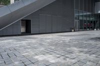 an image of a gray building in the city with a stairs outside the door and on a brick area with the buildings behind it