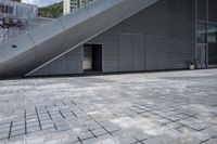 an image of a gray building in the city with a stairs outside the door and on a brick area with the buildings behind it