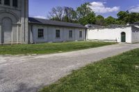 a paved path winds through a grassy area next to an old building with two green doors