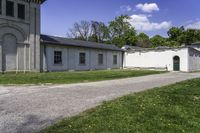 a paved path winds through a grassy area next to an old building with two green doors