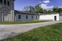 a paved path winds through a grassy area next to an old building with two green doors