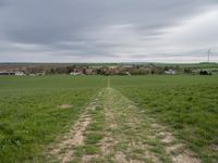 green field with an unpaved path leading to the small town below the horizon