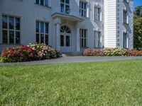 a grass field outside an old white house with a lawn that has flowers and bushes around it