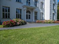 a grass field outside an old white house with a lawn that has flowers and bushes around it