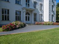 a grass field outside an old white house with a lawn that has flowers and bushes around it