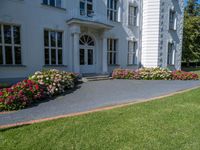 a grass field outside an old white house with a lawn that has flowers and bushes around it