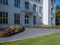 a grass field outside an old white house with a lawn that has flowers and bushes around it
