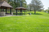 gazebo and benches in open park with grass, trees, and woods behind it