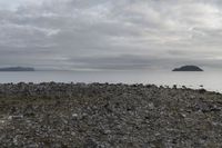 a rocky plain with boulders and water in the distance, with a cloudy sky above the ocean