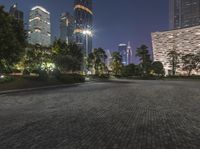 an empty area with a sidewalk and some buildings at night in the background, at a park