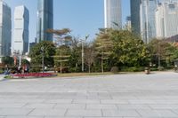 some people on an empty square surrounded by skyscrapers and green trees and shrubs in the foreground