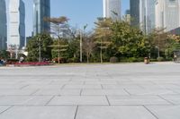 some people on an empty square surrounded by skyscrapers and green trees and shrubs in the foreground