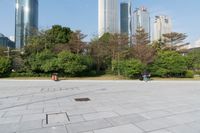 some people on an empty square surrounded by skyscrapers and green trees and shrubs in the foreground