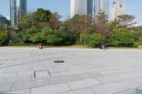 some people on an empty square surrounded by skyscrapers and green trees and shrubs in the foreground