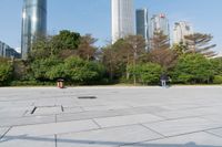 some people on an empty square surrounded by skyscrapers and green trees and shrubs in the foreground